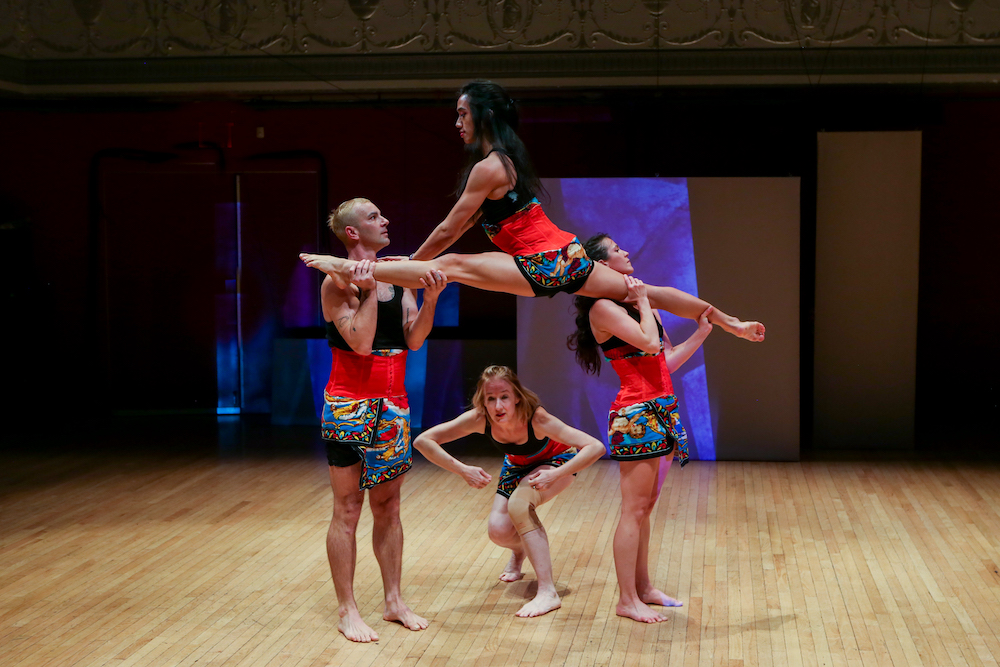 dancers wearing colorful scarves around their waists, lift up one of their group in a split.